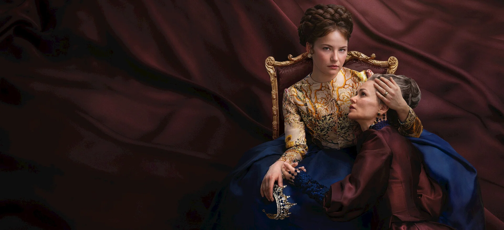 Elisabeth sentada em uma cadeira ornamentada com uma criança ao seu lado, em A Imperatriz.