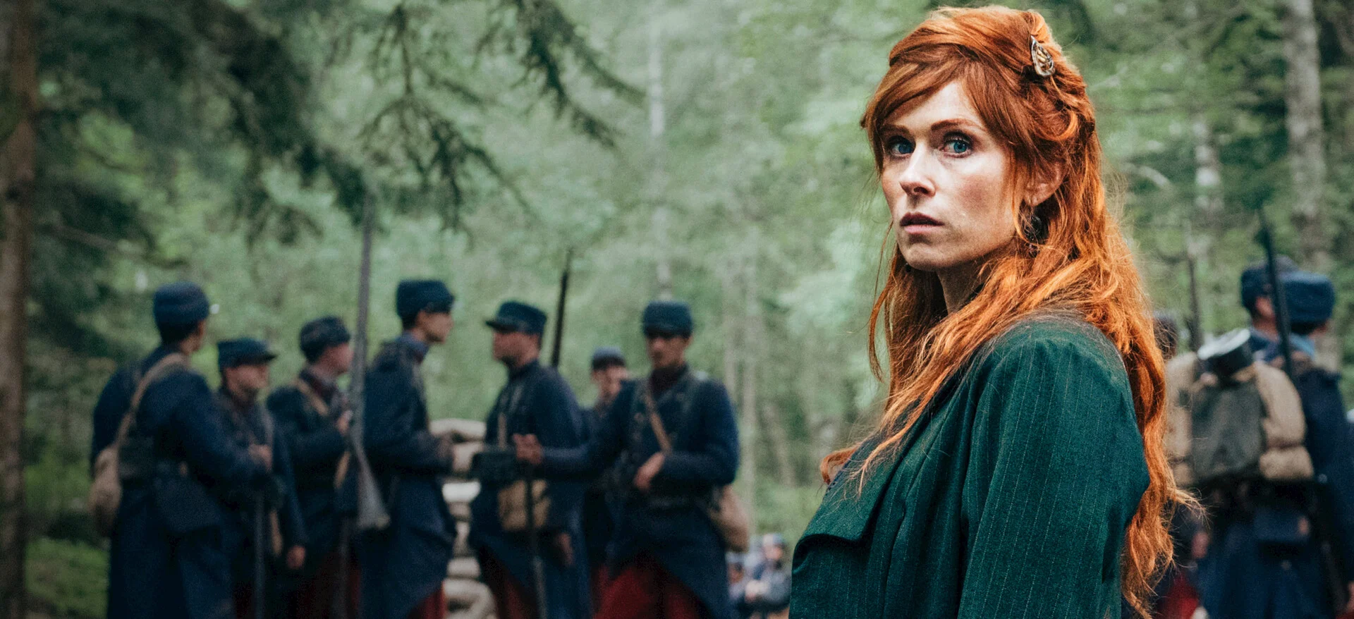 Mulher ruiva com roupa verde olha para trás, com militares ao fundo, em Guerreiras.