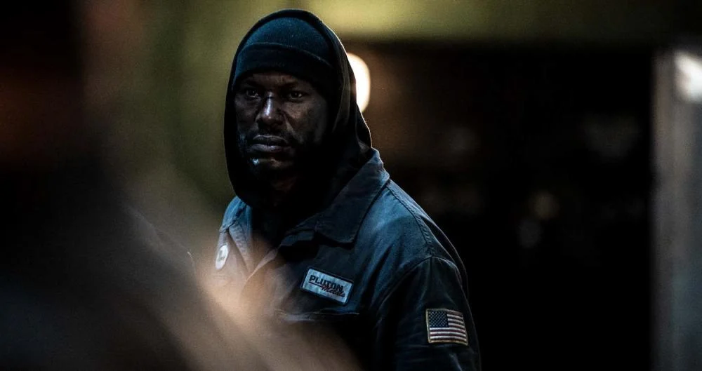 Tyrese Gibson interpreta Mercer, de capuz e uniforme de fábrica, em uma cena escura do filme 1992