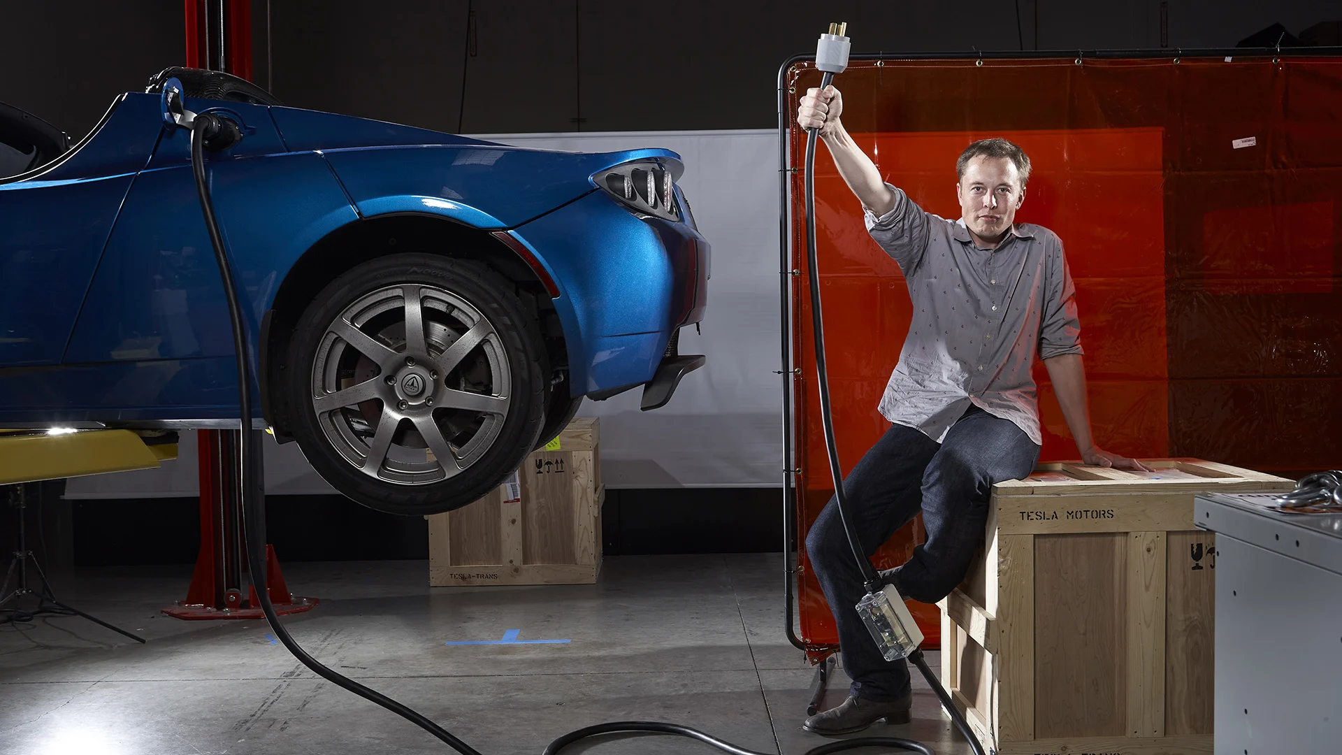 Elon Musk sentado em uma caixa de madeira e segurando um plugue elétrico ao lado de um carro azul elevado