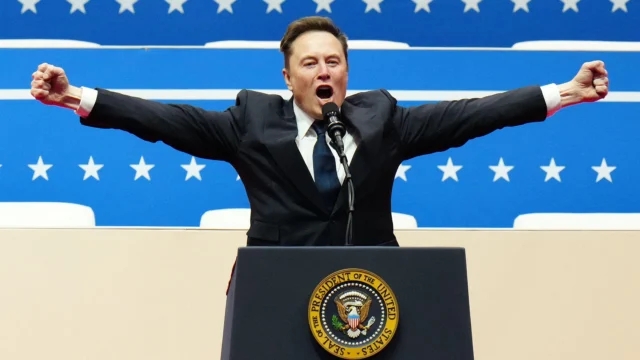 Elon Musk de terno e com os braços abertos diante de um microfone, em imagem de divulgação do documentário Musk