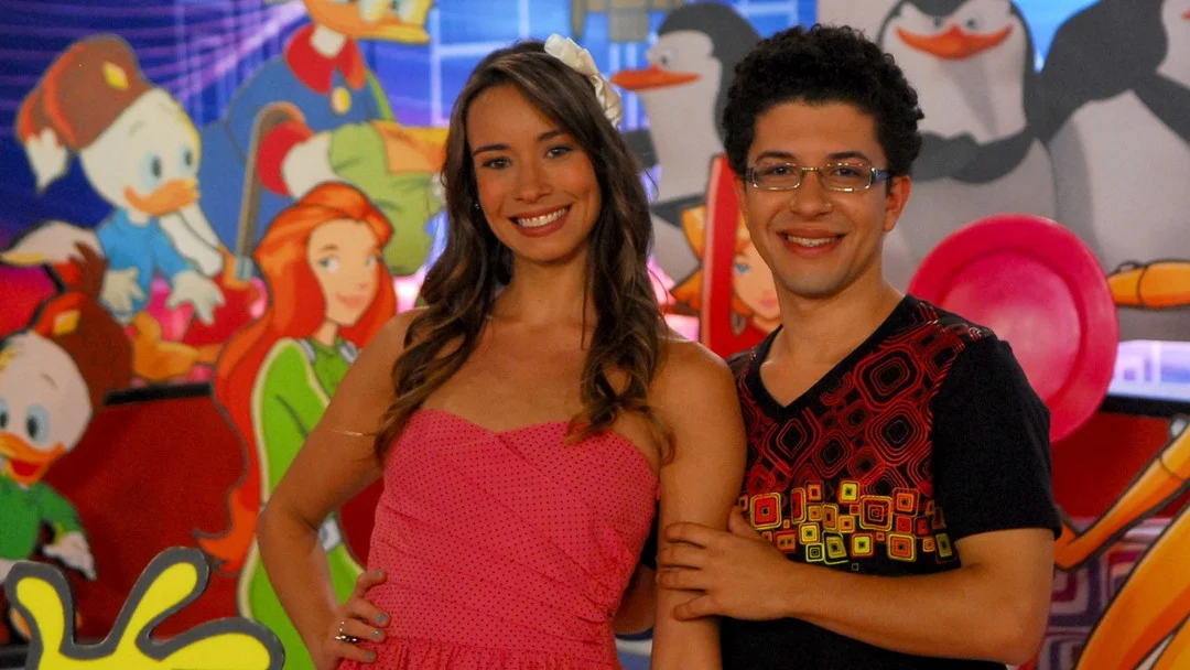 Apresentadores da TV Globinho em cenário antigo com personagens de animações ao fundo