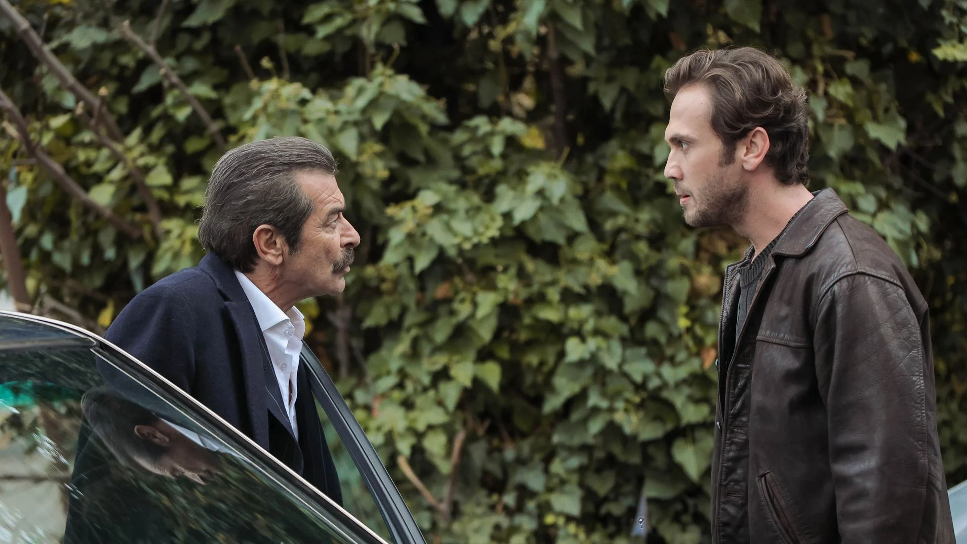 İskender e Devran discutem em uma cena externa da novela turca Deha, perto de um carro