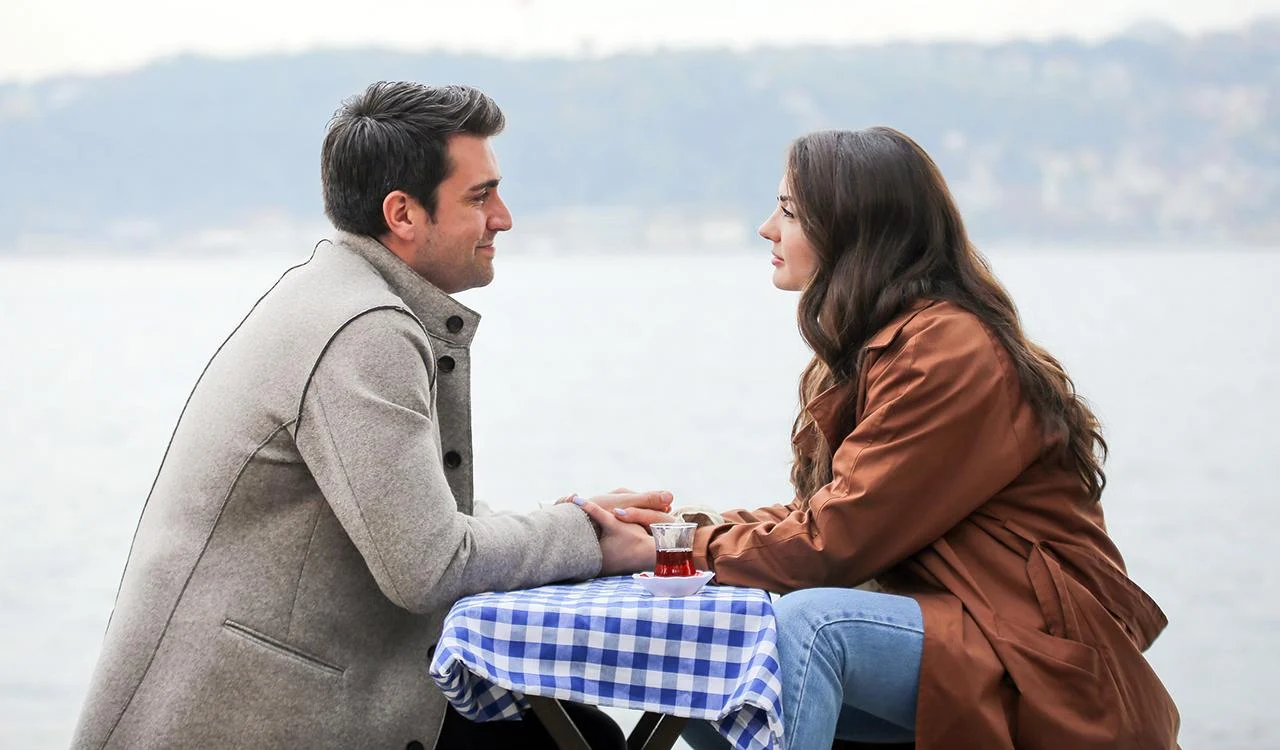 Kerem e Ayşe, personagens de Armadilha do Amor, conversam de mãos dadas a uma mesa perto da água
