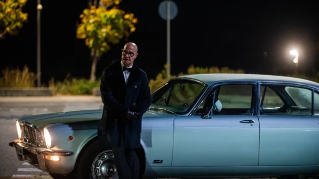 Roger, vivido por Stanley Tucci, em frente a um carro Jaguar durante uma cena noturna de Masterplan