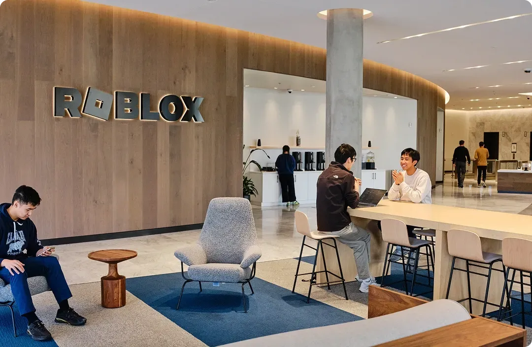 Recepção de um escritório do Roblox, com o nome da empresa na parede e pessoas utilizando dispositivos