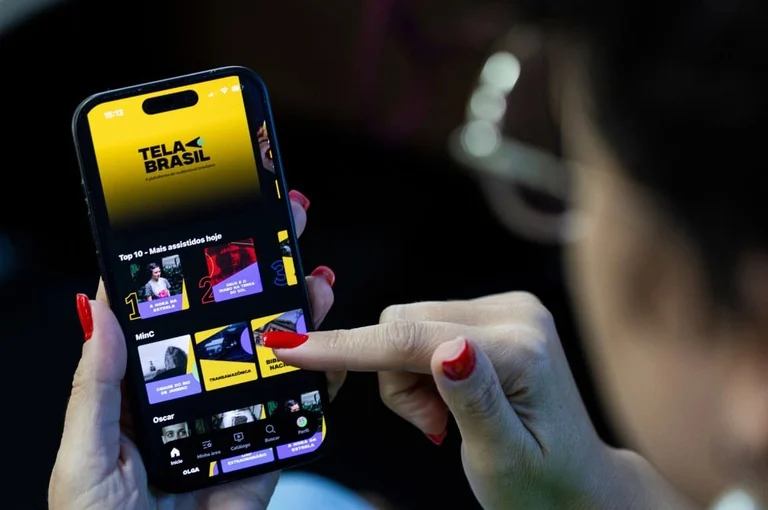 Pessoa segura celular com o aplicativo Tela Brasil aberto e seleciona conteúdo do catálogo