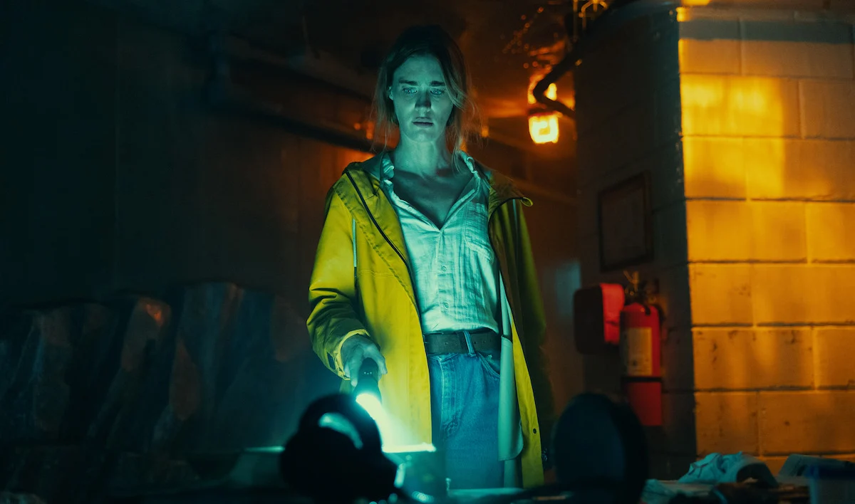Personagem com capa amarela investiga ambiente pouco iluminado em cena da minissérie Profundezas