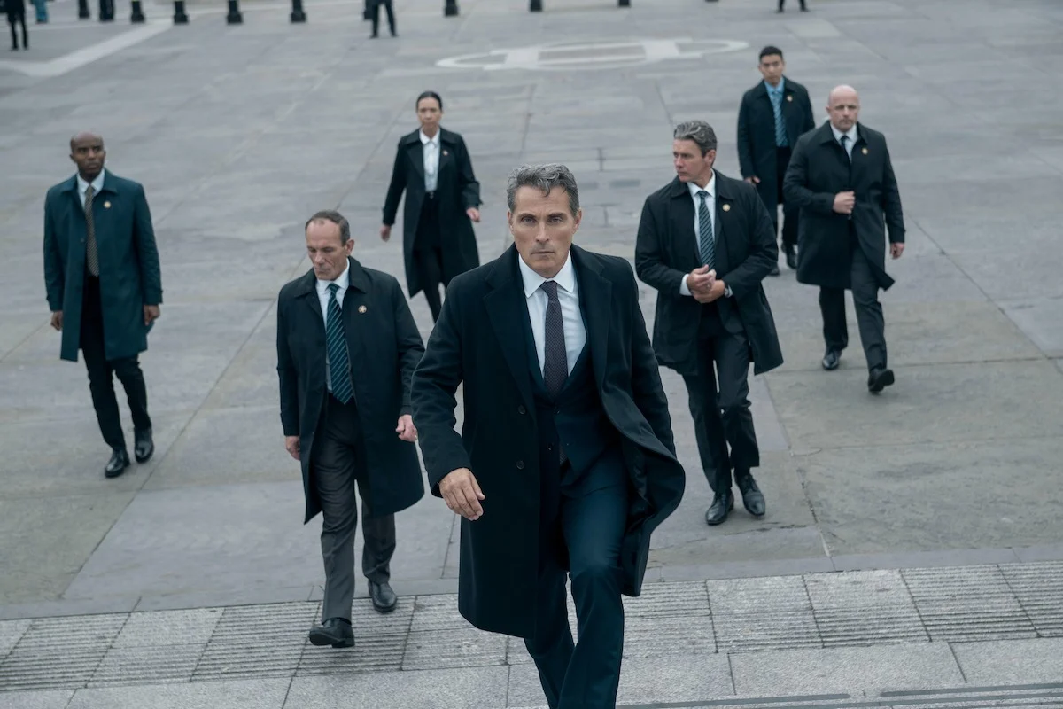 Hal Wyler caminha à frente de uma comitiva de autoridades em A Diplomata