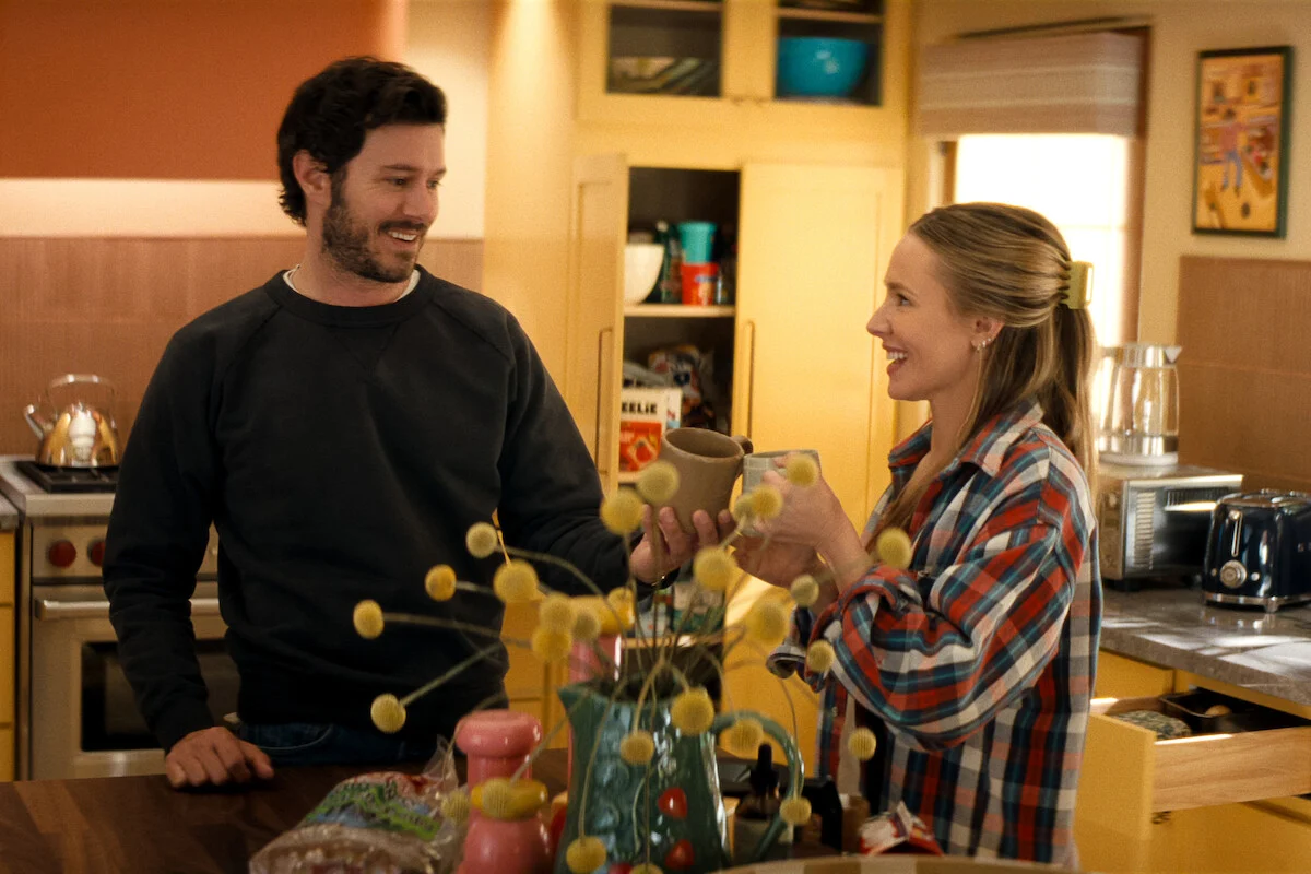 Adam Brody e Kristen Bell como Noah e Joanne conversam segurando canecas em uma cozinha