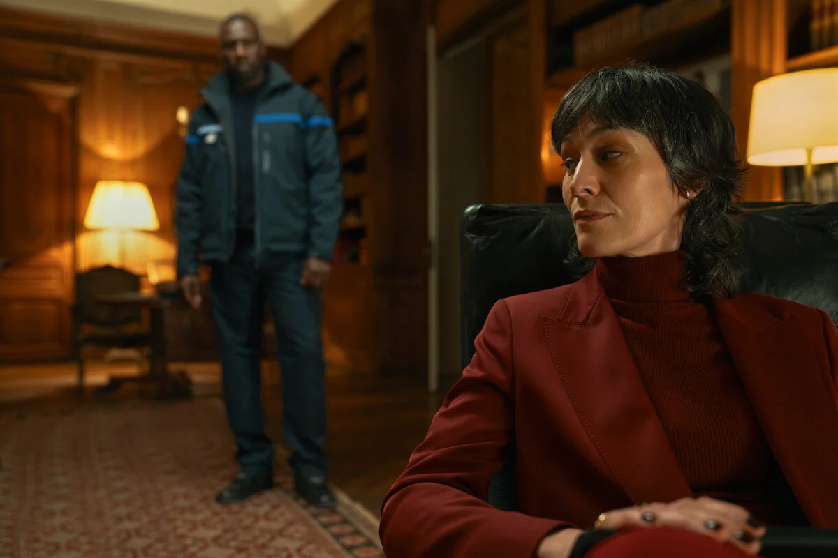 Juliette Pellegrini de paletó vermelho, com Assane ao fundo, em Lupin.