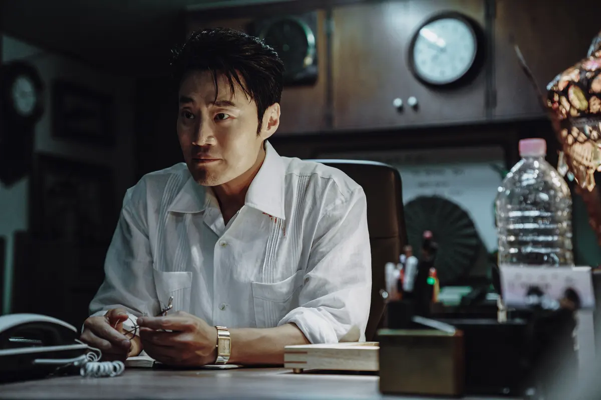 Lee Hee-jun interpreta Mr. S, gerente da agência Spider, sentado à mesa de seu escritório