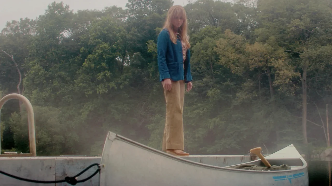 Linda Cardellini como Pamela Voorhees, de jaqueta azul, no cais do lago ao lado de canoas.