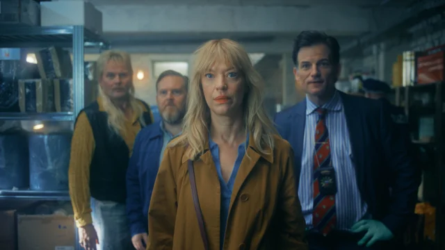 Carlotta Klatt em primeiro plano, com outros personagens atrás dela em uma sala pouco iluminada