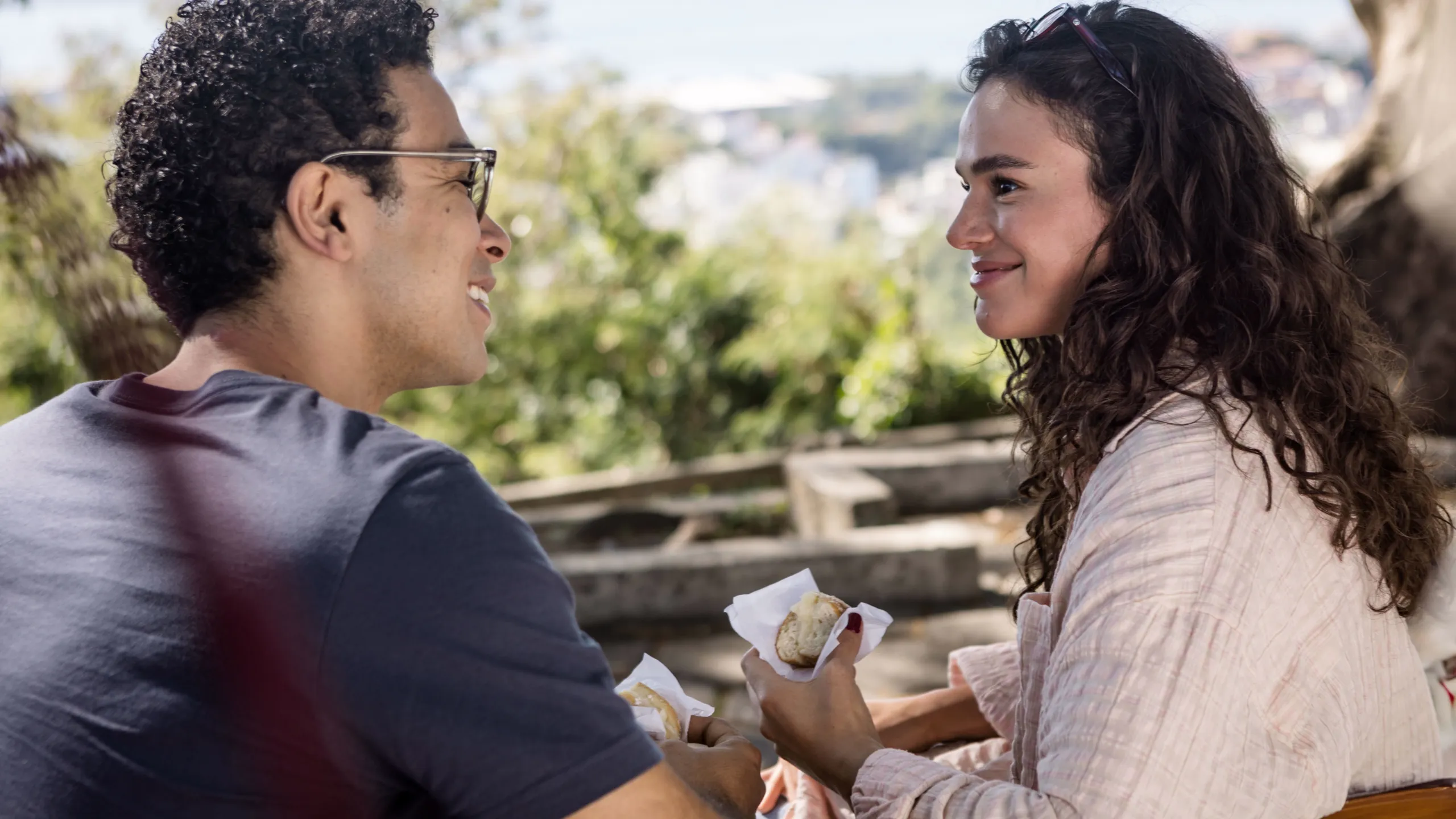 Bia e Victor conversam ao ar livre, sentados e comendo sonhos de padaria em Amor da Minha Vida