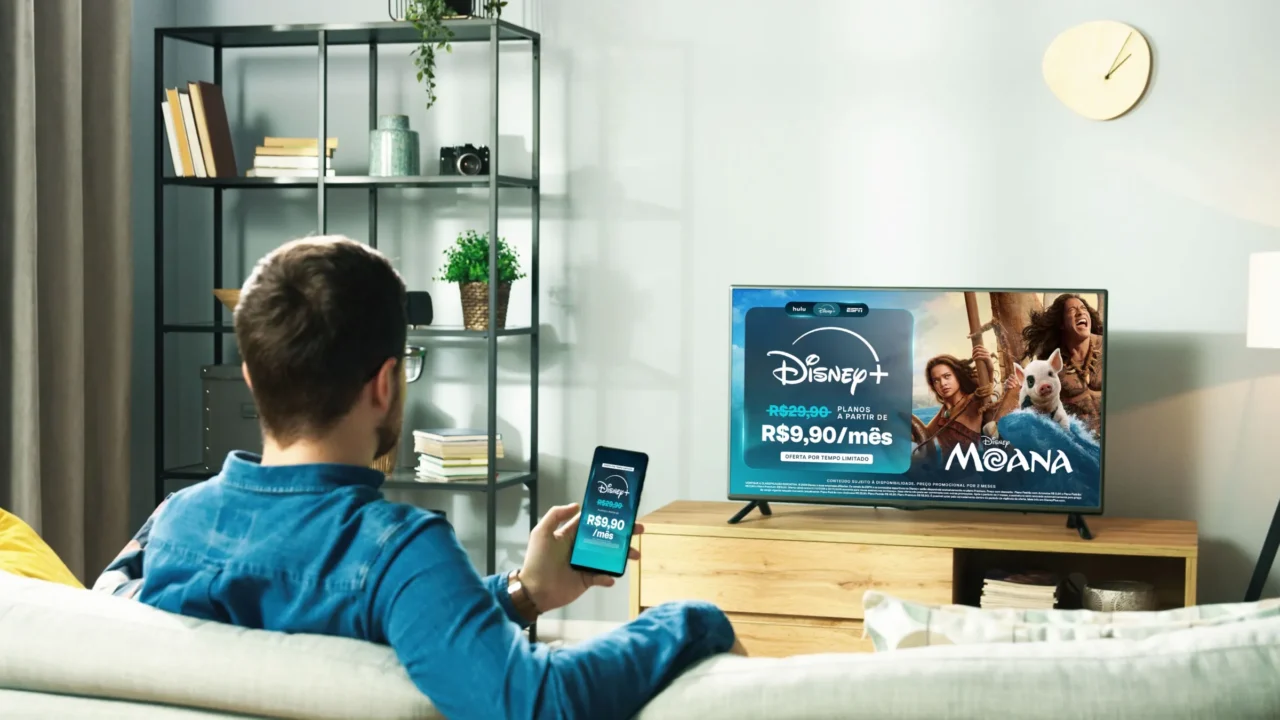 Pessoa acessa o Disney+ pelo celular diante de TV com anúncio da promoção a R$ 9,90 por mês e imagem de Moana