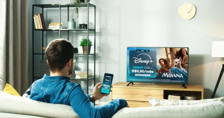 Pessoa acessa o Disney+ pelo celular diante de TV com anúncio da promoção a R$ 9,90 por mês e imagem de Moana