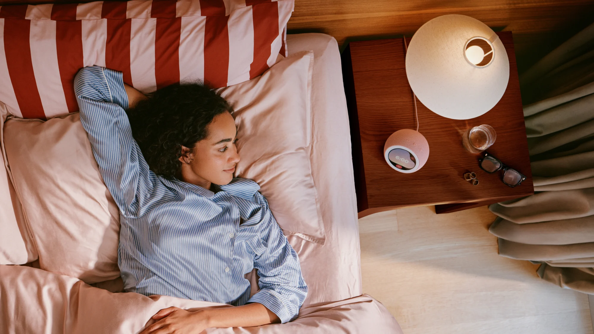 Pessoa deitada na cama com Amazon Echo Time rosa sobre a mesa de cabeceira ao lado de um abajur