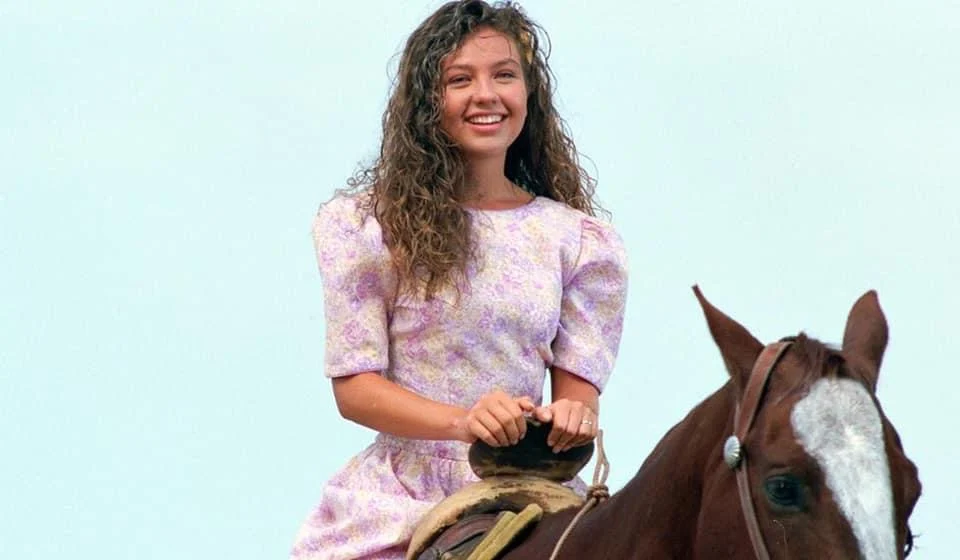 Thalía como Marimar, de vestido floral lilás e cabelo cacheado, montada em um cavalo