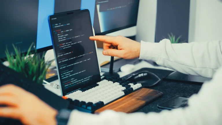 Pessoa programando em notebook ao lado de um monitor com código na tela