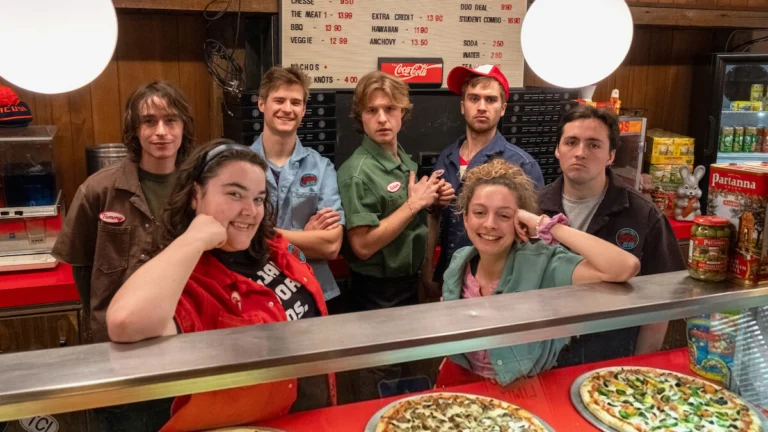 Elenco de Salário Mínimo reunido atrás do balcão de uma pizzaria em imagem de divulgação