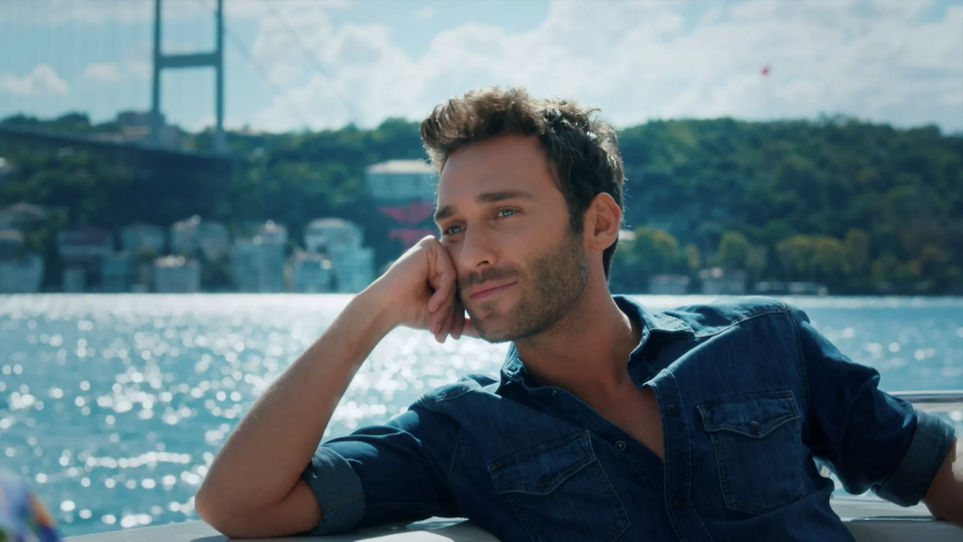 Barış Arduç como Ömer, sentado diante de uma paisagem à beira da água em Amor de Aluguel